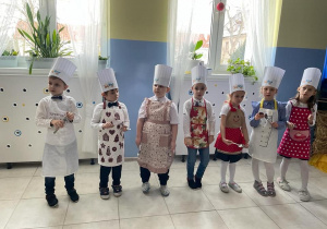 grupa dzieci stoi w szatni, za nimi dekoracja. Dzieci mają założone fartuszki, a na głowach czapki kucharskie, w rękach trzymają różne sprzęty kuchenne